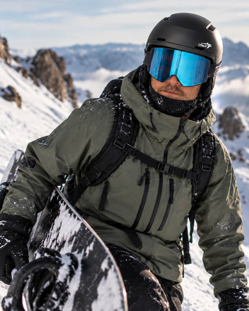 Male freeride snowboarder wearing the lightweight DMN NIVAL matte black snowboard helmet with padded ear protection and blue mirrored ski goggles in the snowy mountains