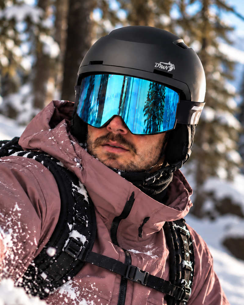 Male snowboarder wearing the DMN NIVAL matte black ski and snowboard helmet with integrated ventilation and blue mirrored ski goggles in a snowy forest